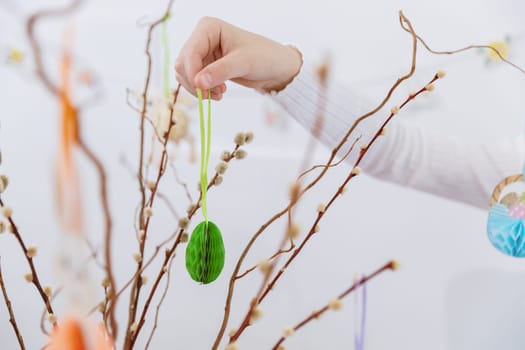 Portrait of one hand unrecognizable Caucasian girl hanging homemade green lace Easter paper egg on pussy willow branches, creating Easter bouquet on spring day, side view very close up selective focus. Concept of happy childhood, happy Easter, children leisure, creative work.