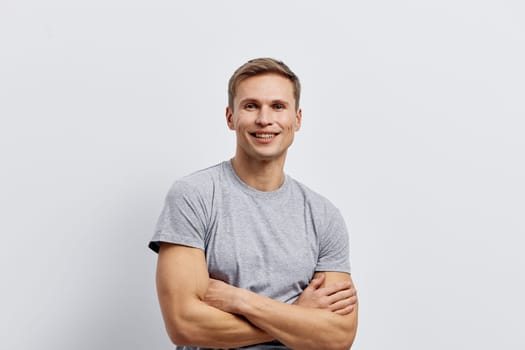 Confident smiling man with arms crossed in casual grey t-shirt standing against a plain white background. Studio image capturing a cheerful, approachable mood with natural lighting, male model, relaxed pose, spring or summer fashion, positive energy.