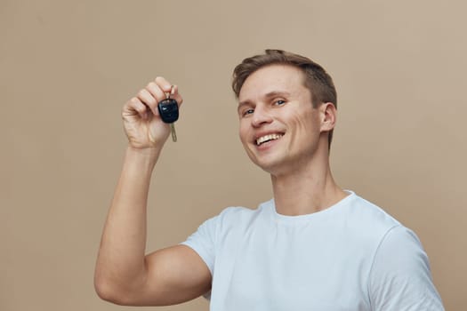 Cheerful young man in a white t-shirt smiling and holding car key, standing against a plain beige background, representing happiness, success, and achievement. Lifestyle and automotive concept