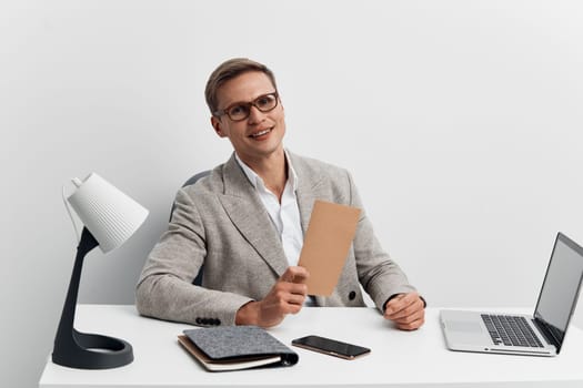 A cheerful professional man dressed in business clothes sitting at a contemporary office desk with a laptop, tablet, and papers, smiling and holding a folder. Bright, clean workspace with a modern professional atmosphere. Concept: workplace success.