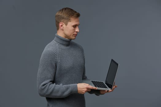 young caucasian man working with laptop using AI chat technology wearing cozy gray sweater isolated on plain gray background showing focus on digital communication and artificial intelligence tools concept