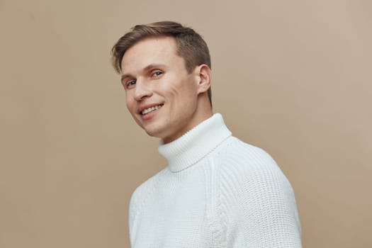 Portrait of a happy young man in a white turtleneck sweater smiling and looking at the camera against a plain beige background. Cheerful and friendly mood, suitable for lifestyle, fashion, or wellness themes.