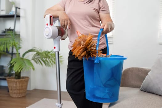 A diligent individual prepares to clean their home, holding a bucket filled with cleaning tools while using a vacuum cleaner, showcasing an organized cleaning routine.