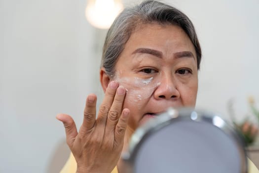A mature woman is dedicated to her skincare routine, applying product with care in front of a mirror, celebrating her beauty and age diversity.