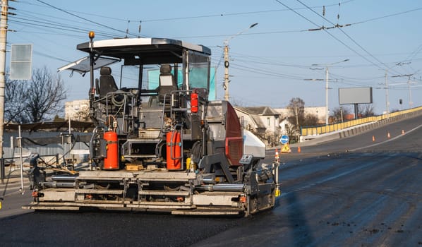 Large asphalt paving machine working on a newly resurfaced urban road. Construction cones and signs visible. The machinery features visible hydraulics and control seats, shot in morning light.