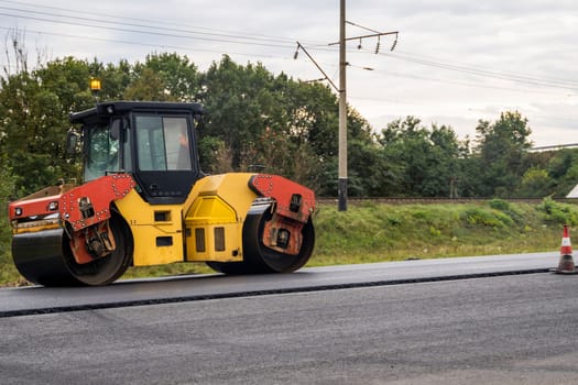 A lone asphalt roller stands beside a construction cone on an active paving site. The background features trees and rail lines, emphasizing road and rail infrastructure development themes.