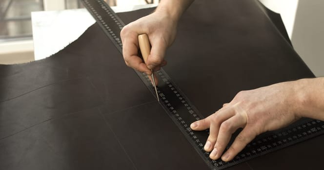 Close-up of a craftsman using a wooden-handled awl and metal ruler to measure and mark straight lines on a dark brown leather hide. The setup highlights precision and traditional leatherworking techniques, set in a naturally lit workspace.
