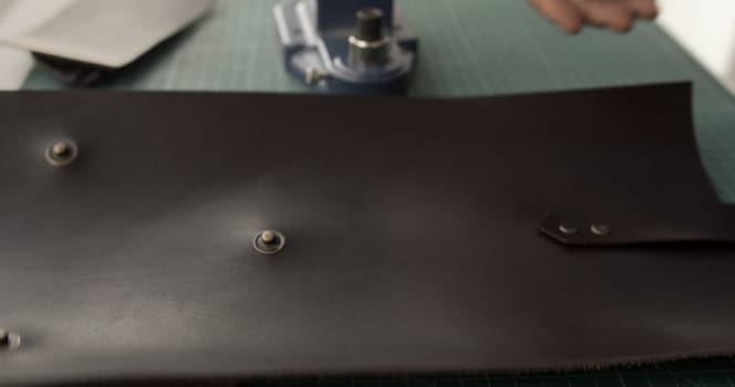 Partially focused image of a leather piece with installed snap fasteners lying on a cutting mat. A hand press machine is visible in the background, hinting at the ongoing assembly process of a handmade item.