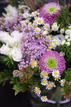 A beautifully arranged bouquet showcases a mix of purple daisies and white roses, complemented by lush greenery and lilac flowers, perfect for a spring celebration or event decor.