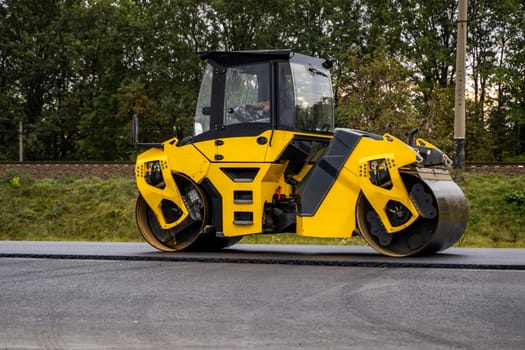 A yellow tandem road roller working on newly laid asphalt, smoothing and compacting the surface. Captured from the side against green trees, emphasizing road construction progress.