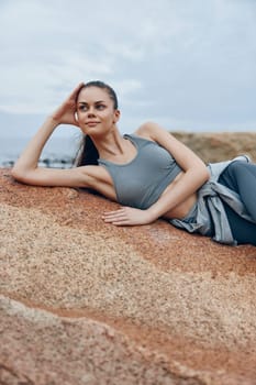 A fit young woman in gray sportswear rests on a rock near the sea, enjoying the calm atmosphere and showcasing a lifestyle of health and wellness