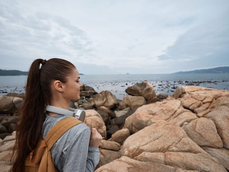 A thoughtful young Asian woman dressed in casual attire stands on rocky terrain, looking towards the ocean, embodying adventure and serenity in a picturesque coastal setting
