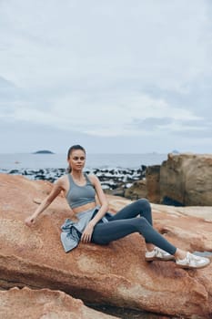 Fit model in gray sportswear poses on rocky beach, reflecting healthy lifestyle and outdoor fitness, evoking calm emotions against a tranquil coastal background
