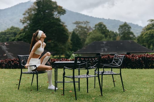 A young woman in fashionable activewear is seated on a chair in a serene outdoor space, exuding calmness and tranquility, surrounded by lush greenery and mountains