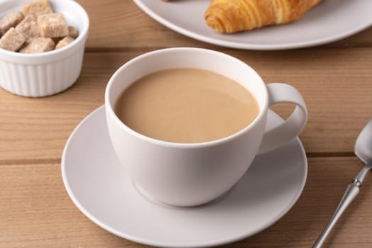 A white cup filled with warm coffee is placed on a saucer next to a croissant and a small bowl of sugar cubes on a wooden table. The setting suggests a cozy breakfast.