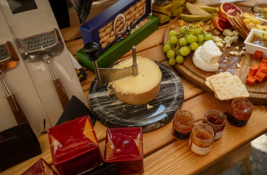 Assorted cheeses on a round wooden board at a farmers market. Camembert cheese, slices of hard cheese, honey, grapes, crackers, bread, grapes, cheese slicers.