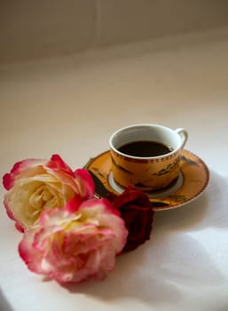 a cup of coffee on a saucer of a soft golden color and delicate pink-red rosebuds nearby. morning coffee in summer