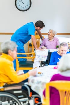Male nurse is taking care of an elderly woman in the dining room of a nursing home while other seniors are eating