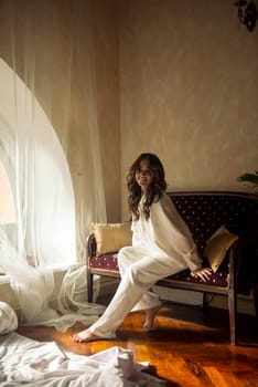 young romantic brunette with long wavy hair sits on the sofa in front of the window. she laughs, dressed in a white silk suit. looks at the camera with a slight smile. vintage furniture to the room