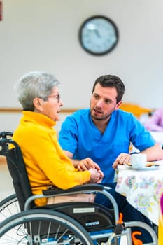 Young male nurse holding hands and comforting elderly disabled woman in wheelchair at nursing home