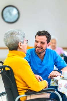 Young male nurse smiling and comforting elderly disabled woman in wheelchair during lunch break at nursing home