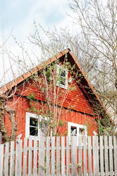 A quaint red house with a triangular roof sits amidst budding trees and shrubs. Wooden fences encircle the property, highlighting the peaceful charm of early spring in a rural setting.