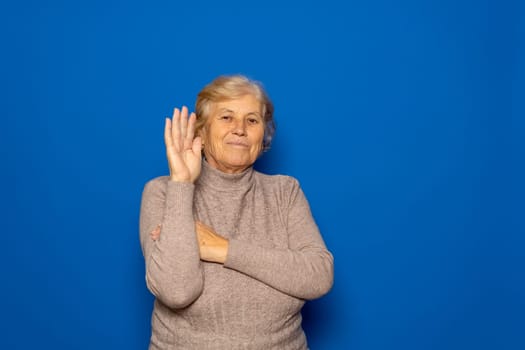 Hello concept. A happy, smiling, elderly Caucasian woman in her 70s waves with her hand outstretched. Isolated on a blue studio background
