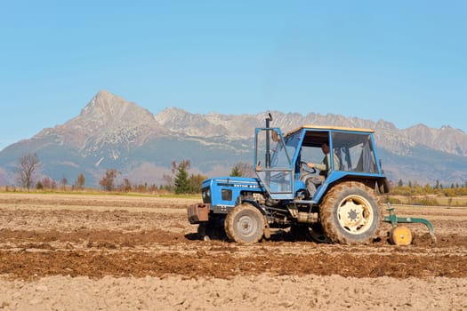Vychodna, Slovakia - October 13th, 2018: Blue tractor plow field, preparing dry ground in autumn. Sunny day, mount Krivan (Slovak symbol) background. Agriculture industry of Liptov region illustration