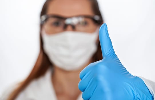 Hand in blue protective glove showing thumb up, blurred woman face with cotton nose mouth mask and protective goggles in background