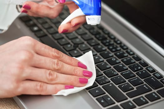 Woman cleaning laptop keyboard with white tissue, detail on her fingers holding paper towel, blue alcohol sprayer near - disinfection concept
