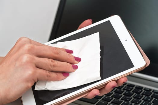 Woman cleaning tablet device with cotton paper tissue, wiping black screen, closeup detail. Blurred laptop keyboard background