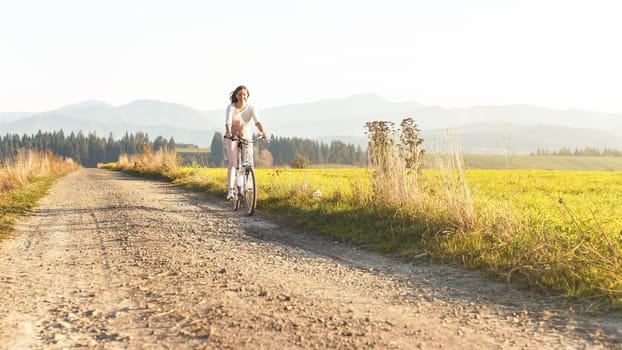 Young woman riding a bicycle on outdoor dusty road, sun lit meadow near