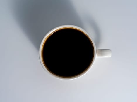 Overhead image of a full white ceramic cup with black coffee casting a long soft shadow on a clean light surface.