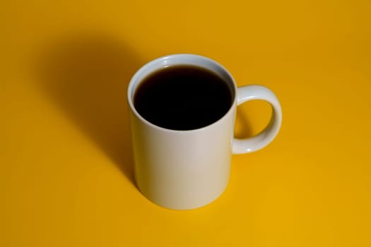 Top side view of a white ceramic mug filled with dark coffee placed on a vibrant yellow surface with dramatic shadow.
