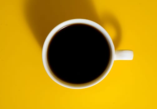 Overhead shot of a white ceramic coffee cup filled with black coffee, placed on a vibrant yellow background with visible shadow.