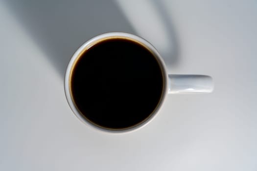 Overhead shot of a white ceramic mug filled with black coffee, placed on a clean light surface with soft natural lighting and visible handle shadow.