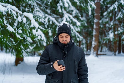 Young man in black winter coat and beanie standing in a snow-covered forest, looking at smartphone with snow on his hat.