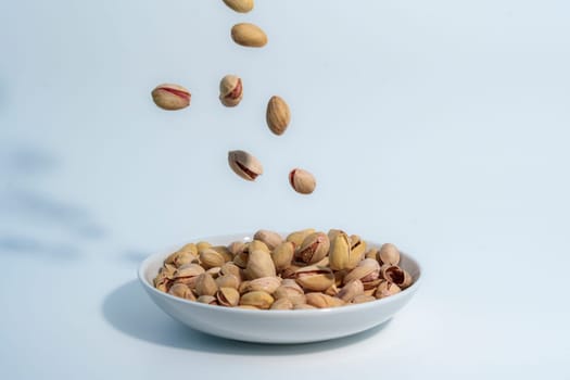 Pistachio nuts falling into a white ceramic bowl already filled with pistachios, captured against a clean light blue background.