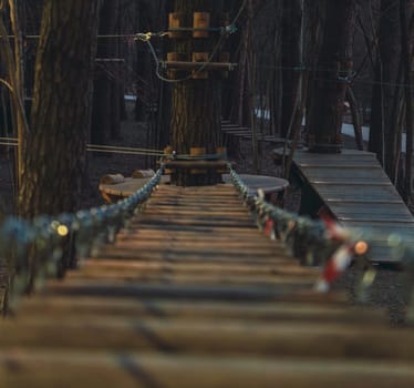 A close-up view of a suspended wooden rope bridge leading to a tree platform in a forest adventure park, captured during sunset in warm light.