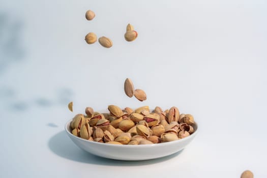 Dynamic image of shelled pistachios falling into a white ceramic bowl, set against a plain white background with scattered nuts and natural shadows.