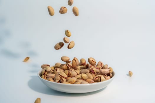 Shelled pistachio nuts captured in motion as they fall into a white bowl already filled with pistachios, against a light-colored background.