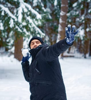 Man in black coat and blue gloves in mid-motion while throwing a snowball in a snow-covered forest during winter