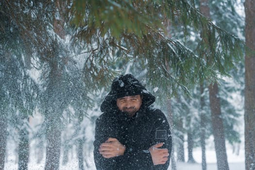 Hooded man in black coat reacts to snow falling from above while standing in snowy forest surrounded by pine trees