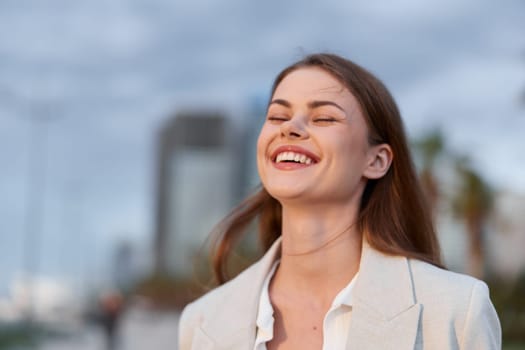 Portrait of a happy young woman smiling outdoors in a casual light blazer, showing natural emotions with blurred cityscape background, perfect for fashion or lifestyle themes.
