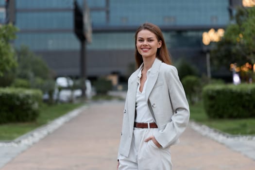 Confident young woman dressed in a light gray business suit stands smiling outdoors in a modern urban park with green foliage, projecting professionalism and modern business style.