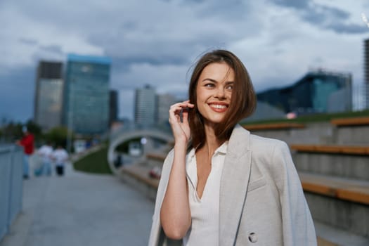 confident woman wearing light beige coat smiling outdoors in urban business environment. Autumn season, stylish casual fashion, modern city background with buildings.