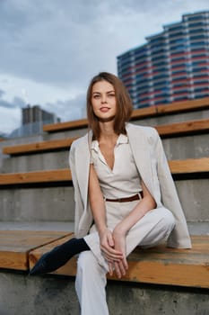 confident woman wearing white business outfit and beige blazer sits on wooden stairs in urban environment with modern architecture and cloudy sky, expressing professionalism and calmness