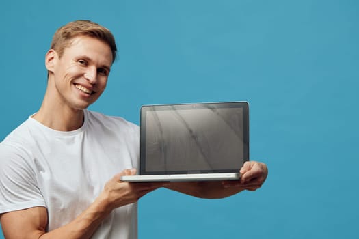 smiling man holding laptop wearing casual white t-shirt on blue background with confident friendly expression representing digital nomad lifestyle and remote work concept using technology and internet connectivity. Concept: digital nomad