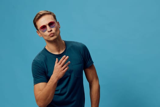 Confident young man with trendy sunglasses and casual blue T-shirt standing in front of a solid light blue backdrop. Male model with cool style, relaxed pose, and fashionable appearance, conveying youthful confidence. Concept: modern fashion.