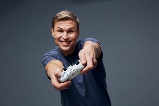 Happy young man with short brown hair and blue casual shirt holding a game controller, smiling enthusiastically while looking at the camera against a plain gray background, capturing excitement and engagement, lifestyle and gaming concept